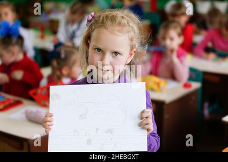 A preschool girl or primary school student holds a sheet of paper with a child's drawing in fr Stock Photo