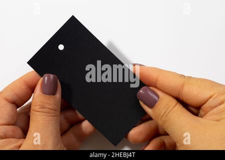 Two hands with cardboard black rectangular blank tag with little hole in upper part for clothes in center of white background. Tag mock up. Copy space Stock Photo