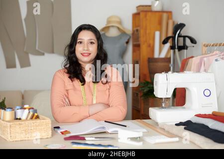 Portrait of smiling female tailor sitting at table with sewing machine, basket of threads, fabrics and notepad Stock Photo