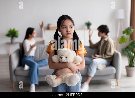 Lonely sad chinese teen girl with toy suffers from scandal of parents, young asian lady and guy quarreling at home Stock Photo