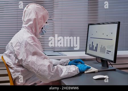 Doctor in protective suit sitting at his workplace and typing on computer keyboard, he working with diagrams at office Stock Photo