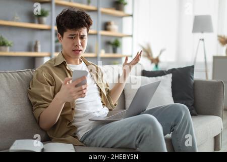 Confused asian man looking at mobile phone using laptop Stock Photo