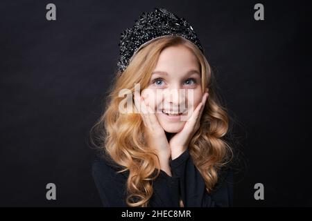 Children's portrait cute girl with crown on her head folded her hands near her face in surprise. Happy posing on a black background in the studio. Beautiful long wavy blonde hair. Open blue eyes Stock Photo