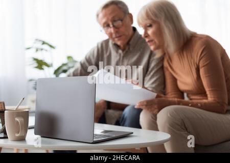 Senior Couple Reading Papers Using Laptop Sitting On Couch Indoors Stock Photo