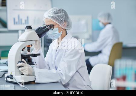 Young doctor in mask and lab coat looking through the microscope at the table, she examining medical samples Stock Photo
