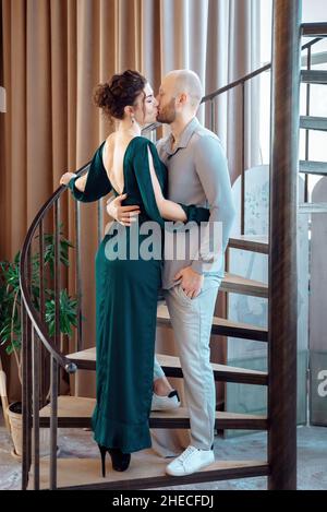 Lovely gently couple embracing with closed eyes standing on a spiral staircase. Passionate young man and woman are kissing. Attractive brunette woman in long evening dress and stylish bearded man. Stock Photo