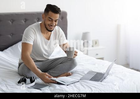 Remote Work. Arab Freelancer Man Working With Laptop And Notepad In Bed Stock Photo