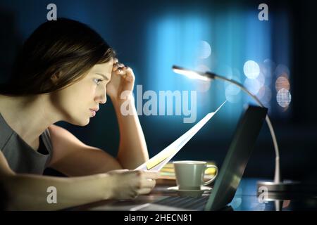 Side view portrait of a worried businesswoman checking document online with a laptop in the night Stock Photo