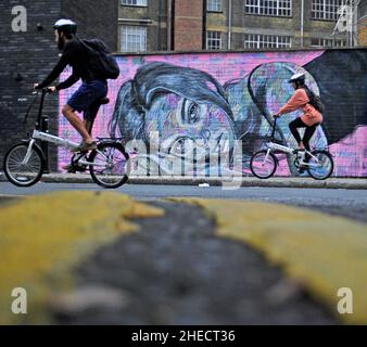 A low pov take on a London Street art portrait, which makes it look like the figure is resting on the pavement. Also in shot are two cyclists. Stock Photo