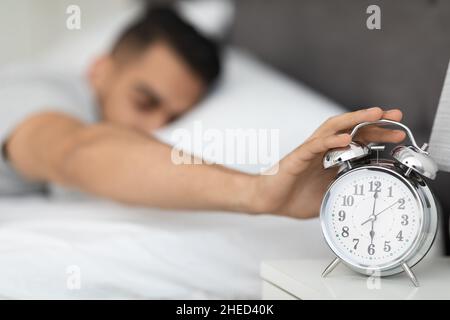 Young Arab Man Reaching Alarm Clock On Bedside Table With Hand Stock Photo