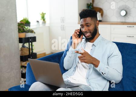 Handsome bearded African-American guy in casual jeans shirt talking on the smartphone and using laptop sitting on the sofa in cozy living room, positive man has pleasant phone conversation Stock Photo