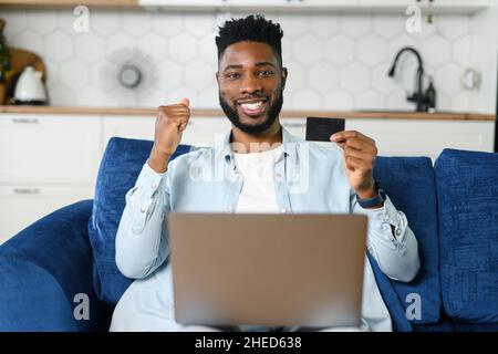 Overjoyed African man holds banking card and laptop on the laps looks at camera, making order online, excited guy using app for booking, paying online for long awaited purchase with credit card Stock Photo
