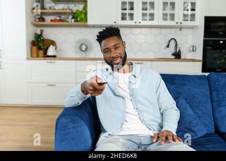 Serene and carefree African-American guy holding TV remote controller watching television movie, man relaxed sitting on the comfortable sofa at living room, points the remote at the camera