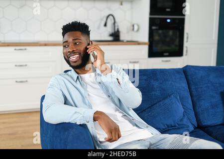 Happy handsome African-American guy has fun phone conversation sitting on the couch at home, overjoyed biracial man talking on the smartphone, chatting on the mobile phone, feels happy and joy Stock Photo