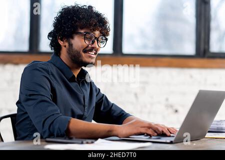 Portrait of dark-haired man using laptop in home office. Curly male entrepreneur looking at the screen and smiling toothy, windows on the background Stock Photo
