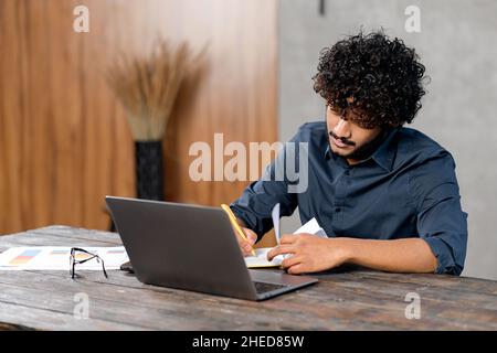 Online training. Young guy student with curly hair studying at home while sitting in the modern room and using laptop. He writing with a pen in a notebook Stock Photo