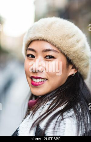 Delighted Chinese female in warm clothes and beret looking at camera with smile while standing on street against blurred background