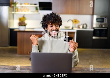 Smiling Indian man holding banking card and laptop, making purchases or ordering online. Excited guy paying online with credit card while sitting on the table and rejoicing Stock Photo