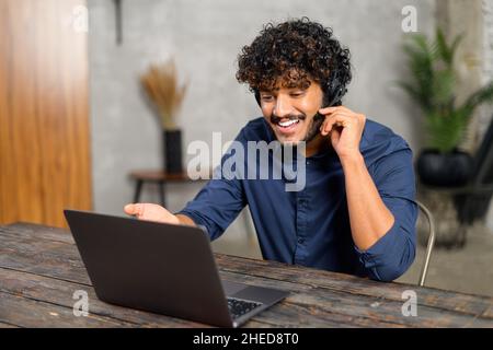 Focused young businessman wearing headphones, holding video call with clients on laptop. Concentrated Indian man giving online educational class lecture, consulting customer Stock Photo