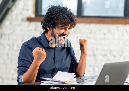 Excited Indian man sitting at the table, looking at the laptop screen and rejoicing at good news. Surprised emotional lucky man with wide smile Stock Photo