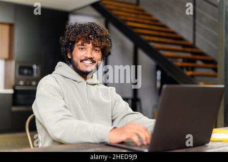 Handsome young freelancer man with beard sitting at the table and looking at the camera, taking a break from working on laptop, dreaming about vacation or salary, enjoying working from home Stock Photo