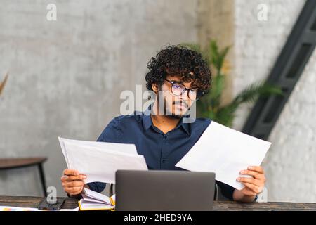 Executive Indian male office employee doing paperwork while sitting at the desk with laptop in office. Focused and serious Hindu man looking through sheets of documents, preparing report Stock Photo
