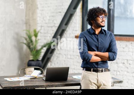 Smiling bearded Indian businessman standing near desk and looking away. Young positive male student with modern flat on background. Proud and successful ethnic small business owner Stock Photo