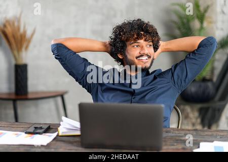 Unshaved man sitting at the table and smiling, while feeling comfort and relaxation at his workplace. Confident office worker sitting in front of the laptop and looking away Stock Photo