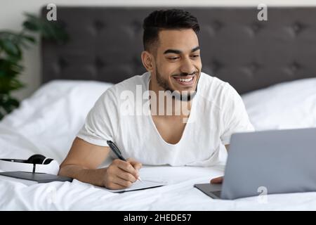 Online Education Concept. Arab Man Studying With Laptop While Lying In Bed Stock Photo