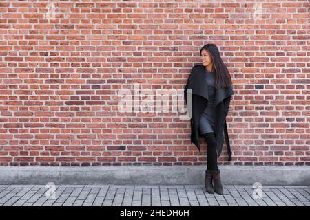 Asian young woman fashion model standing on street against brick wall background wearing winter wool coat and elegant dress for work Stock Photo