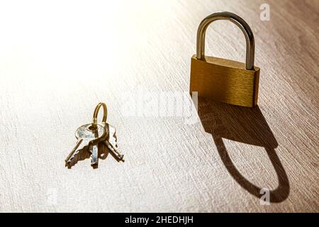 On a wooden table there is a padlock planted and next to it a ring with three keys. The light comes intense from above and casts a hard, elongated sha Stock Photo