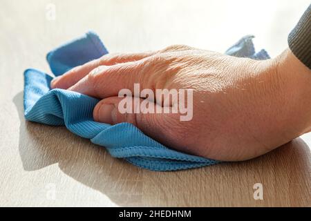 An older man's hand is wiping a wooden table with a blue cloth cloth. The light comes intense from the front and casts the shadow on the tabletop. Stock Photo