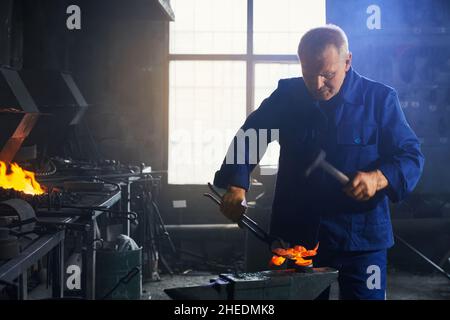 Front view of senior man in dark blue work uniform creating beautiful decorations for modern fence in smithy. Concept of process making different details for metal or fence. Stock Photo