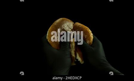 Close up of hand in black cooking gloves breaking bread into two pieces isolated on black background. Food and cooking concept, fresh burger bun being Stock Photo