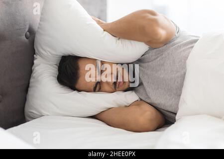 Insomnia Concept. Annoyed Middle Eastern Guy Covering Ears With Pillow Stock Photo