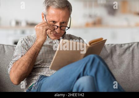 Senior Man With Poor Eyesight Reading Book Above Eyeglasses Indoor Stock Photo