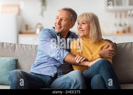 Happy Mature Spouses Hugging Looking Aside Sitting On Couch Indoor Stock Photo