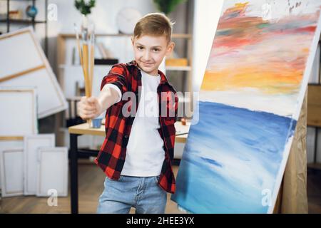 Happy caucasian schoolboy in casual clothes standing near easel and holding multiple paint brushes. Cute boy enjoying painting process art modern creative school. Stock Photo