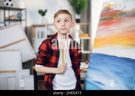 Thoughtful caucasian boy in checkered shirt looking aside while searching for inspiration at art studio. Little student standing near canvas and holding paint brushes. Stock Photo