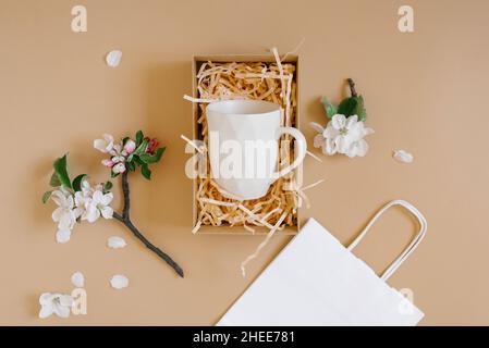 An open gift box with peach-colored paper shavings containing a creative trendy mug with a geometric pattern, a white paper bag and apple flowers on a Stock Photo
