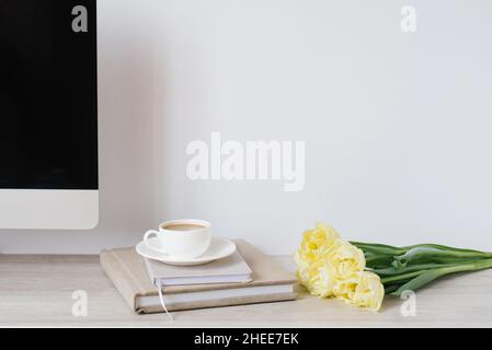 Spring home office still life composition. Blank laptop screen. Yellow tulips in a vase, a cup of coffee and notepads on the table. Office workplace. Stock Photo