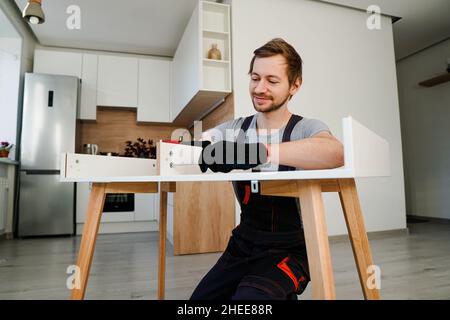 Smiling busy man in construction uniform assembles table with screwdriver, work with pleasure in living room. Stock Photo