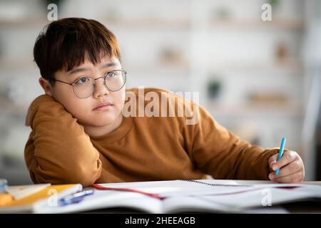 Korean schooler chubby boy dreaming about something while doing homework Stock Photo