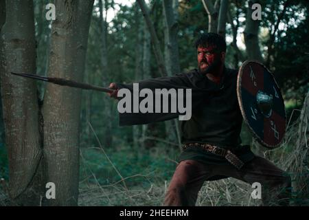 Angry male viking with face in blood fighting with spear and shield at medieval historic reenactment in woodland Stock Photo