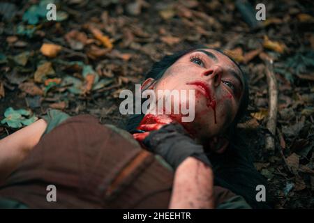 High angle of dying bleeding female viking holding neck while lying on ground during reconstructed medieval fight in forest Stock Photo