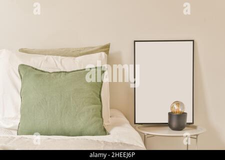 Pillows on comfortable bed near lamp and blank frame on bedside table near white wall in bedroom Stock Photo