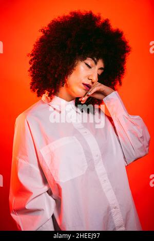 Self assured young Hispanic female model in white shirt touching face with eyes closed in studio with bright red neon illumination Stock Photo