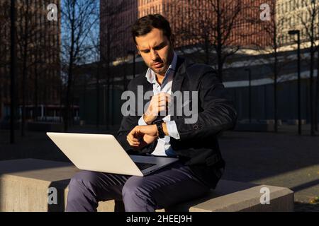 Confident male entrepreneur checking time on wristwatch while browsing netbook on stone border during online work on sunny street in city Stock Photo