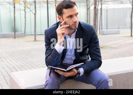 Thoughtful male entrepreneur in formal wear looking away with pensive face while sitting with notepad on stone barrier in city Stock Photo