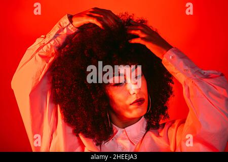 Self assured young Hispanic female model in white shirt touching curly dark hair and looking again in studio with bright red neon illumination Stock Photo